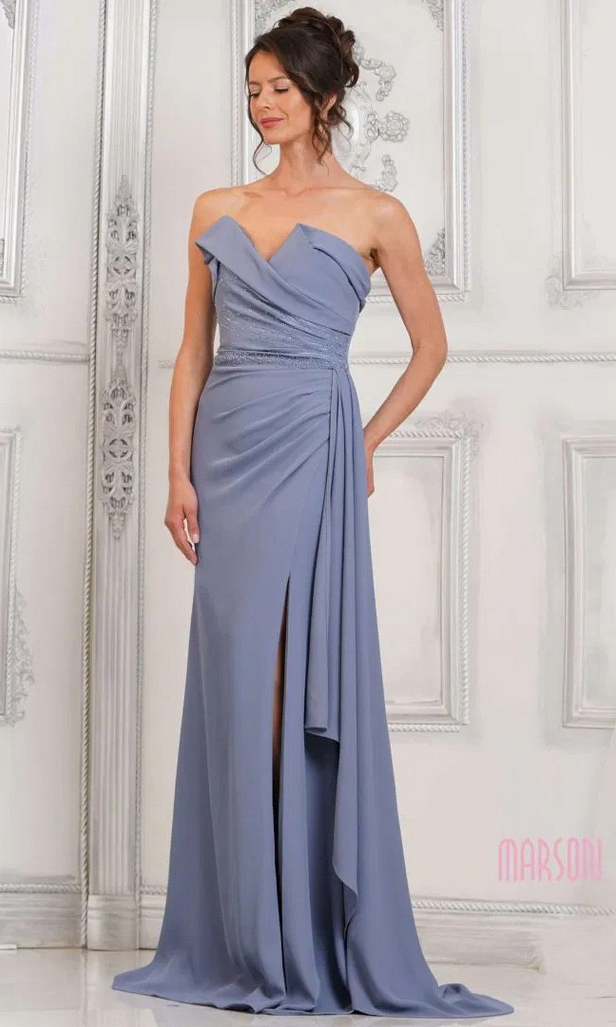 A woman poses before an ornate white wall wearing the Marsoni MV1304 long formal dress in slate blue, featuring a strapless beaded bodice, a draped floor-length skirt with thigh-high slit.